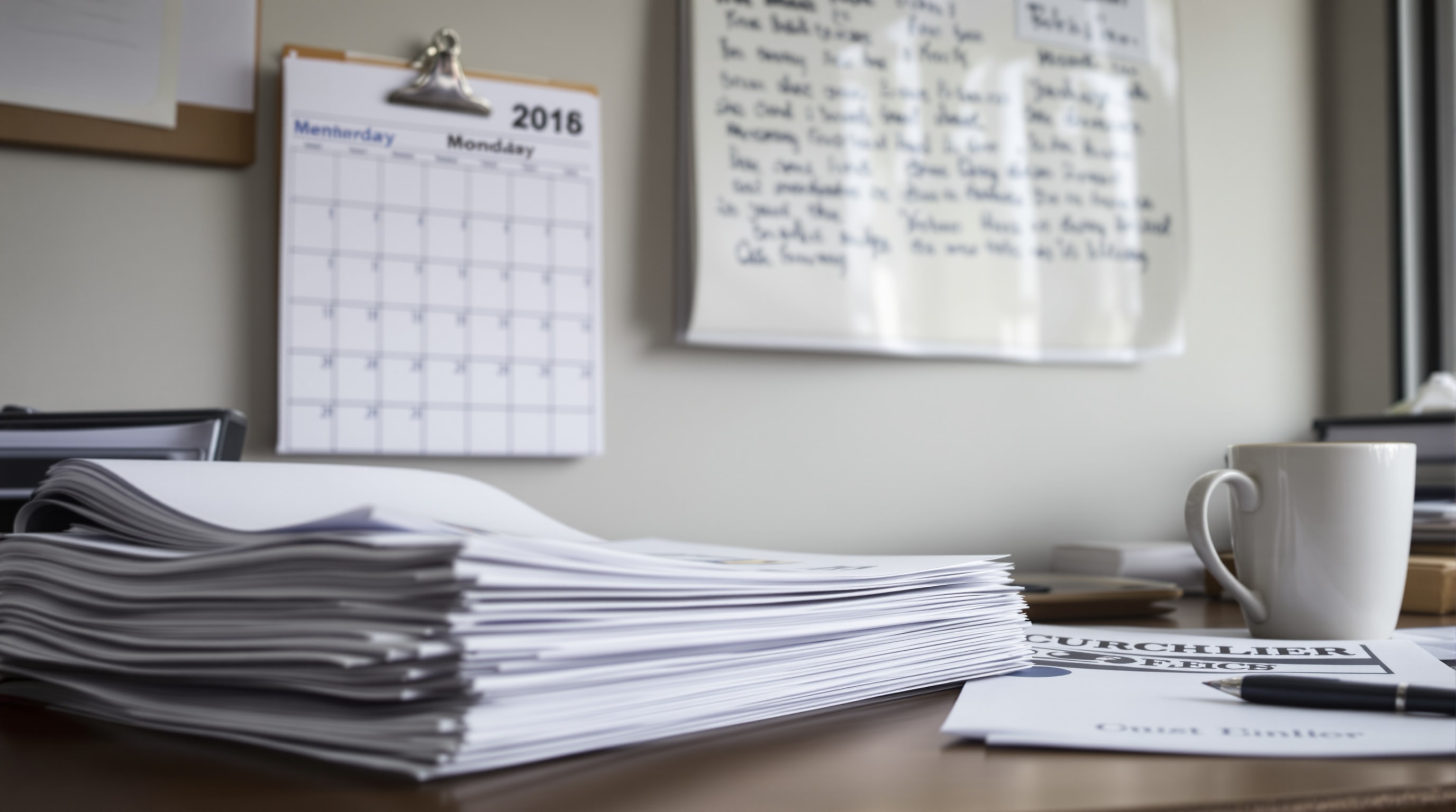A church office desk with folded paper bulletins, a newsletter and a wall calendar behind